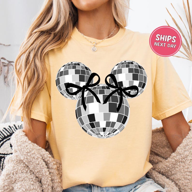 A woman wearing a yellow t-shirt with a graphic of a disco ball and two black bows.