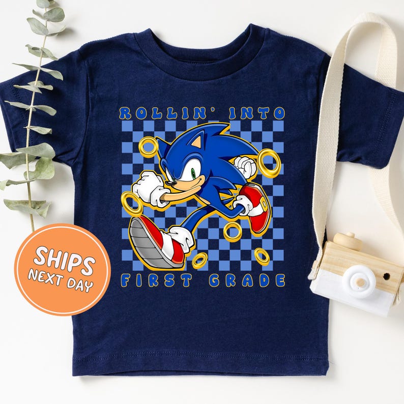 A navy blue t-shirt with a graphic of Sonic the Hedgehog, a popular video game character, printed on it. The t-shirt is displayed on a white background, along with a camera and a plant.