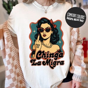 A white t-shirt with a graphic design featuring a woman with sunglasses and a red bow in her hair, along with the text "Chinga La Migra" and the word "FUCK ICE" written below.