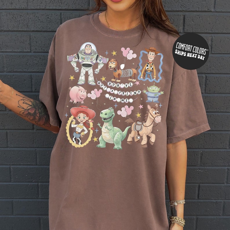 A person wearing a brown t-shirt with a graphic design featuring characters from the Toy Story franchise, including Buzz Lightyear, Woody, and other beloved characters.