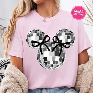 A woman wearing a pink t-shirt with a graphic of a disco ball and two black bows.