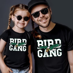 May include: Two people wearing matching black t-shirts with the text "BIRD GANG" in white block letters and a green and white eagle graphic. Both are smiling and wearing sunglasses.