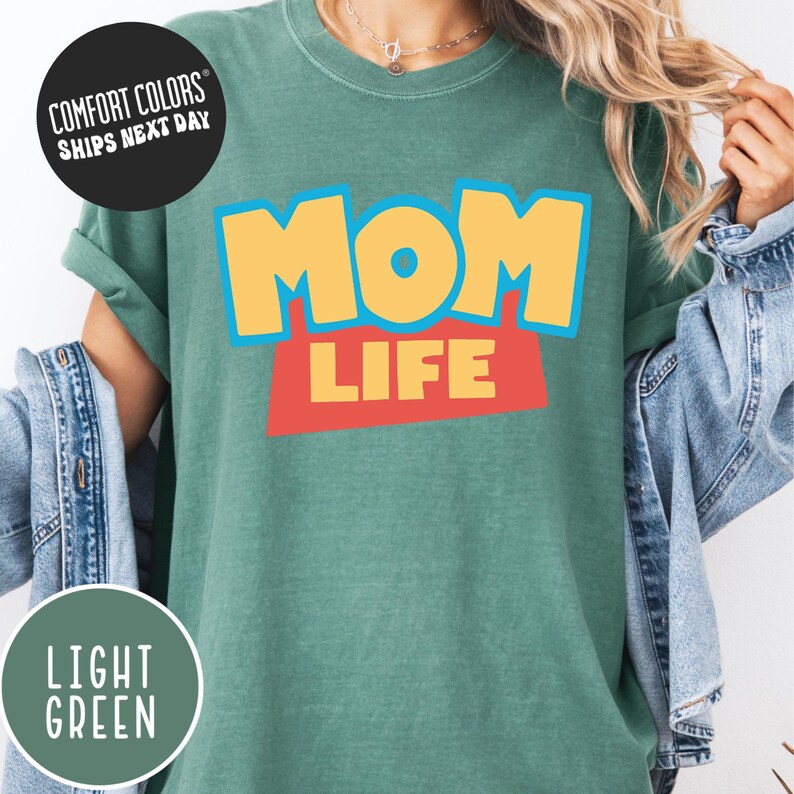 A person wearing a green t-shirt with the text "MOM LIFE" printed on it, along with a denim jacket.