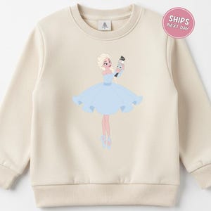 May include: Cream-colored sweatshirt featuring a ballerina illustration. The ballerina is depicted in a light blue dress, holding a nutcracker. The design includes ballet shoes and a light blue bow. The sweatshirt has a crew neck and long sleeves.