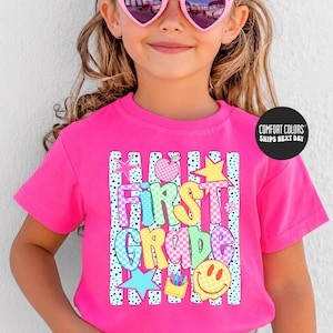 May include: A bright pink short-sleeved t-shirt featuring a graphic print with the text "FIRST GRADE" in colorful checkered letters. The design includes a pink apple, a yellow star, a blue star, and a yellow smiley face with a crayon box.