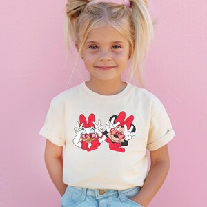 A young girl with blonde hair is smiling and wearing a white t-shirt with a graphic of Minnie Mouse and Mickey Mouse.