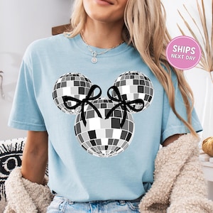 A woman wearing a light blue t-shirt with a graphic of a disco ball and two black bows.
