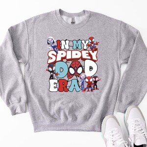 A gray sweatshirt with a graphic design featuring various Spider-Man characters and the text "IN MY SPIDERY DAD ERA" printed on it.