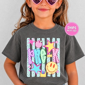May include: A charcoal gray t-shirt featuring a graphic print with the text PRE-K in colorful checkered letters. The design includes a pink apple, a yellow star, a blue star, a yellow smiley face, and a small yellow container of crayons.