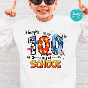 A young boy wearing a white sweatshirt with a colorful graphic design celebrating the 100th day of school.