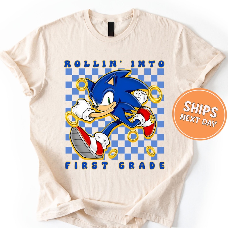 A white t-shirt with a graphic design featuring a cartoon character, likely Sonic the Hedgehog, and the text "Rollin' into first grade" printed on it.