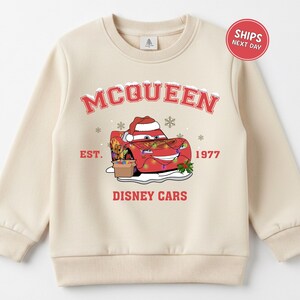 A beige sweater with a graphic design featuring a red car character from the Disney movie Cars, along with snowflakes and text.