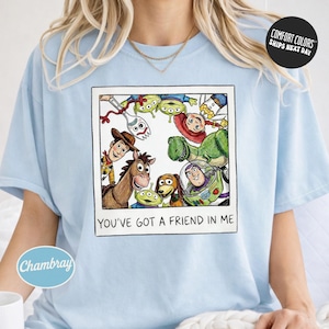 A person wearing a light blue t-shirt with a graphic design featuring characters from the Toy Story franchise, including Woody, Buzz Lightyear, and other beloved characters.
