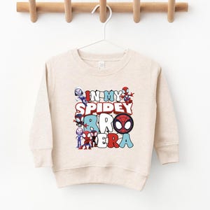 A beige sweater with a graphic design featuring Spider-Man characters and the text "IN MY SPIDER BABY" printed on it.