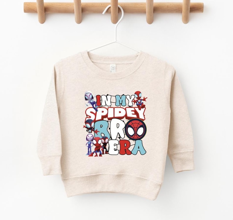 A beige sweater with a graphic design featuring Spider-Man characters and the text "IN MY SPIDER BABY" printed on it.