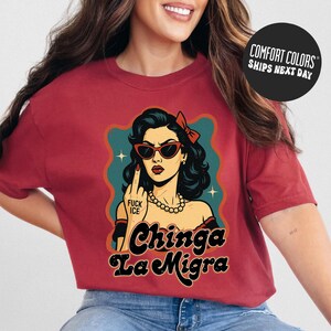 A woman wearing a red t-shirt with a graphic design featuring a woman with sunglasses and the text "Chinga La Migra" (Spanish for "I'm not a chink").