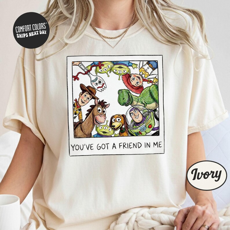 A person wearing a white t-shirt with a graphic design featuring characters from the Toy Story movie, including Woody, Buzz Lightyear, and other beloved characters. The text below the image reads "You've Got a Friend in Me".