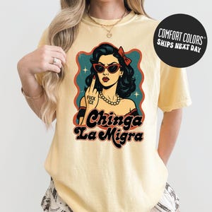 A woman wearing a yellow t-shirt with a graphic design featuring a woman with sunglasses and the text "Chinga La Migra" (Spanish for "chung ching" or "chung ching").