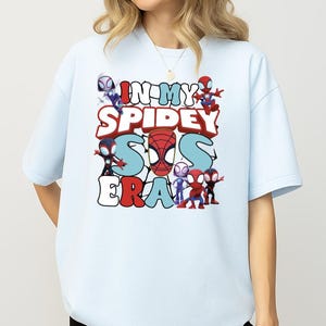 A person wearing a white t-shirt with a graphic design featuring various Spider-Man characters and text.