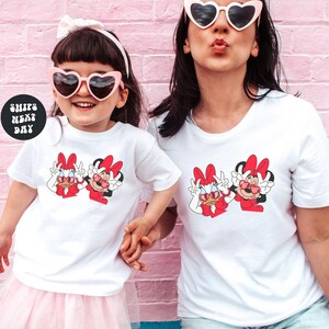 Two women wearing matching white t-shirts with cartoon Minnie Mouse characters are posing together against a pink brick wall.