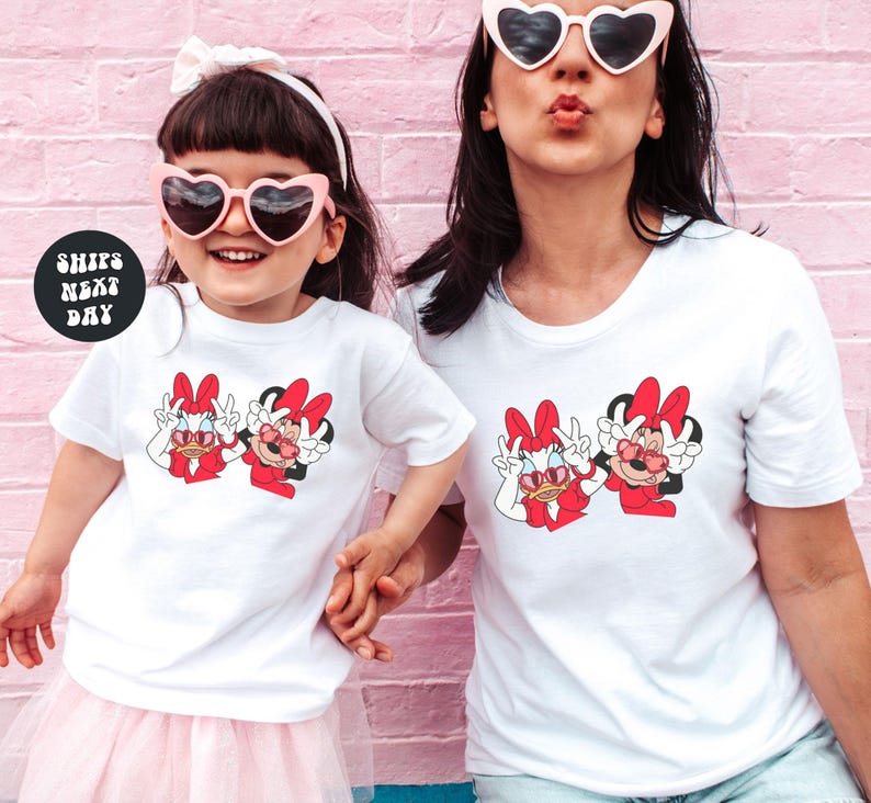 Two women wearing matching white t-shirts with cartoon Minnie Mouse characters are posing together against a pink brick wall.