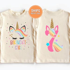 May include: Two cream-colored children's t-shirts. One features a unicorn face with a rainbow mane, gold horn, and the text "BIRTHDAY GIRL." The other shirt displays a pink unicorn with a rainbow mane and the number 7.