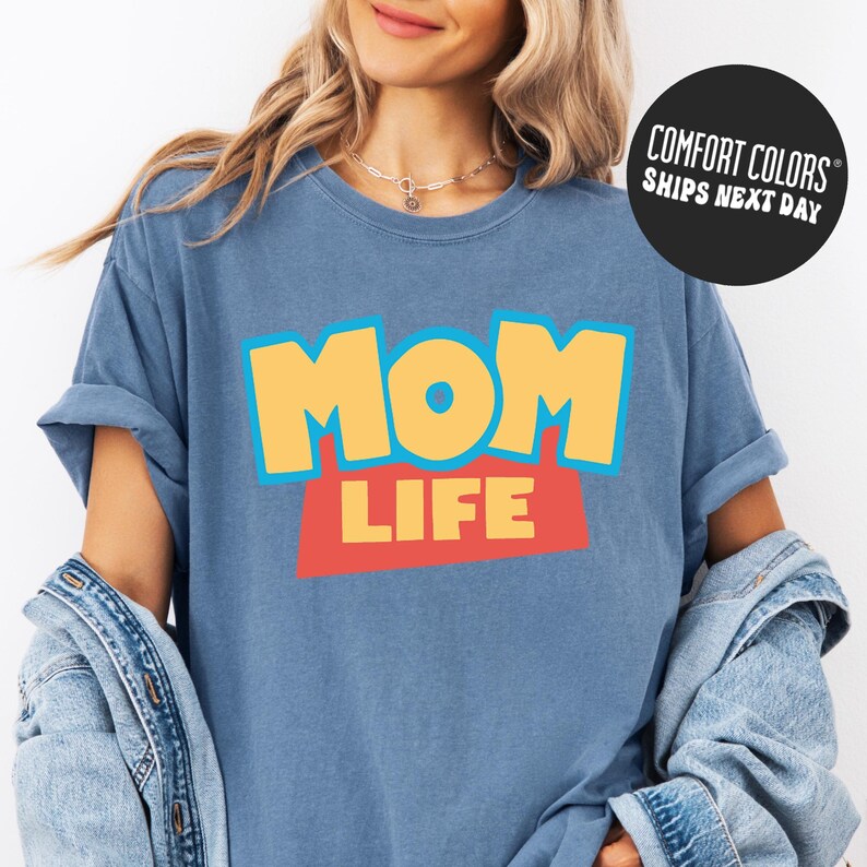 A woman wearing a blue t-shirt with the text "MOM LIFE" printed on it, along with a denim jacket.