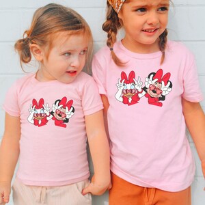 Two young girls wearing matching pink t-shirts with Minnie Mouse designs stand together against a white wall.