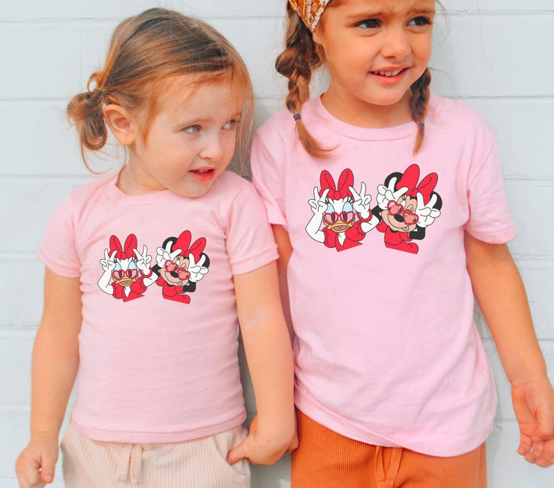 Two young girls wearing matching pink t-shirts with Minnie Mouse designs stand together against a white wall.