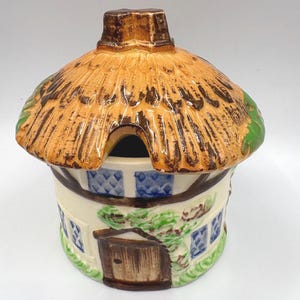 May include: A ceramic cottage-shaped jar with a thatched roof and a small chimney. The jar has a cream-colored base with blue window details and a brown door. Green vines and leaves are painted around the door.