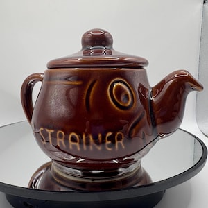 Puede incluir: Tetera de cerámica marrón con tapa y boquilla, con un diseño en forma de pez. La palabra "STRAINER" está inscrita en el lateral. La tetera está sobre una superficie reflectante.