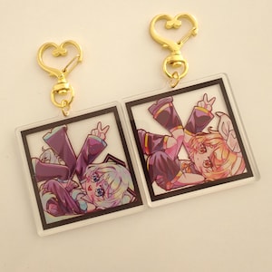 Peut inclure: Deux porte-clés de style anime avec des fermoirs dorés en forme de cœur. Chaque porte-clés présente un charm carré en acrylique transparent avec un personnage de dessin animé en vêtements violets et roses, faisant le signe de la paix. Le fond est beige clair.