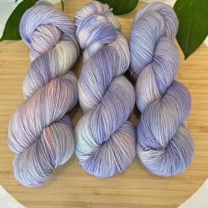 May include: Three skeins of yarn in shades of lavender, white, and pink sit on a wooden surface. The yarn is arranged in a row, with each skein showing a gradient of colours. Green leaves are visible in the background.