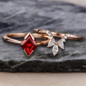 Puede incluir: Dos anillos de oro rosa. Uno presenta una gema roja en forma de diamante. El segundo es una banda curva con pequeñas gemas transparentes. Los anillos se muestran sobre una superficie oscura y texturizada. El nombre de la marca "GalaxyJewelriesIN" está en la imagen.