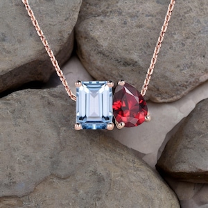 May include: A rose gold necklace featuring two gemstones: a rectangular light blue aquamarine and a pear-shaped red garnet. The gemstones are set in rose gold prongs and connected by a delicate chain. The necklace is displayed on a bed of gray rocks.