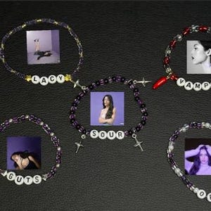 Può includere: Cinque braccialetti di perline con diversi design. I braccialetti sono viola, neri e rossi, con perline di lettere bianche che compongono parole come "LACY", "SOUR", "GUTS", "VAMPIRE" e "OR". Ogni braccialetto ha una piccola immagine allegata.