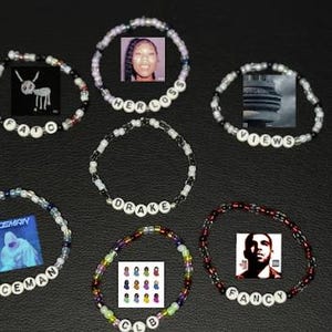 Könnte beinhalten: Eine Sammlung von Armbändern mit Perlen und verschiedenen Designs und Texten. Einige Armbänder zeigen Albumcover oder Bilder, während andere Wörter wie "FATD", "HER LOSS", "VIEWS", "ICEMAN", "CLB", "DRAKE" und "FANCY" mit Buchstabenperlen buchstabieren.