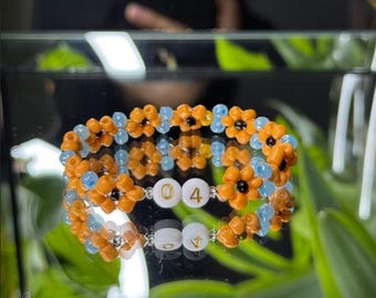 May include: A beaded bracelet featuring orange flower-shaped beads with black centers and light blue beads. The bracelet includes white beads with the numbers "04" and the letter "A". The bracelet is displayed on a reflective surface.