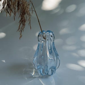 Könnte beinhalten: Eine hellblaue Glasvase mit gewelltem Rand und vertikaler Rippenstruktur. Die Vase hat einen bauchigen Körper, der sich nach oben verjüngt, und enthält einen einzelnen getrockneten braunen Grashalm mit federartigen Samenständen, der nach links ragt.