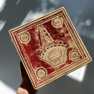 Puede incluir: Azulejo cerámico cuadrado con base crema y diseño rojo. El diseño incluye una estructura que se asemeja a una torre minera, un barco lleno de material y emblemas circulares. El azulejo tiene un borde de pequeños puntos.