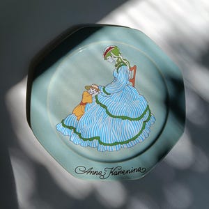 Puede incluir: Plato octogonal azul claro con una ilustración de una mujer con un vestido a rayas azules y un niño. El plato tiene un borde verde y las palabras "Anna Karenina" en cursiva.