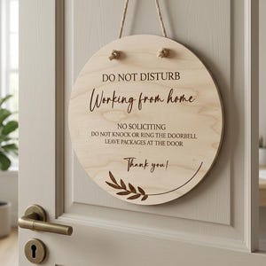 May include: A round light-colored wooden door sign hanging from a gold hook by a jute rope. The sign features black text reading DO NOT DISTURB, Working from home, NO SOLICITING, DO NOT KNOCK OR RING THE DOORBELL, LEAVE PACKAGES AT THE DOOR, Thank you!