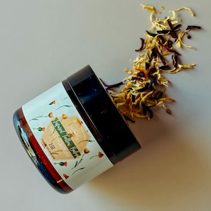 May include: A jar of Infused Tallow Balm with Hibiscus and Calendula, 2 oz. The jar has a black lid and a white label with floral illustrations. Dried yellow and red flower petals are scattered around the jar.