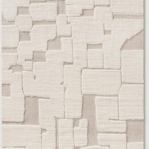 Puede incluir: Una alfombra rectangular de color blanquecino con un patr&oacute;n geom&eacute;trico texturizado. El dise&ntilde;o presenta formas irregulares en relieve que se asemejan a bloques o piezas de rompecabezas, creando una superficie t&aacute;ctil. La paleta de colores neutros y el dise&ntilde;o abstracto la hacen adecuada para varios estilos de interior.