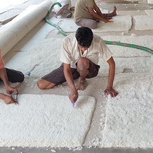 Puede incluir: Escena de taller donde se fabrica una alfombra grande de color blanquecino. Dos personas est&aacute;n arrodilladas, usando herramientas para trabajar en la superficie de la alfombra. Tambi&eacute;n se ve una alfombra enrollada y una manguera verde.