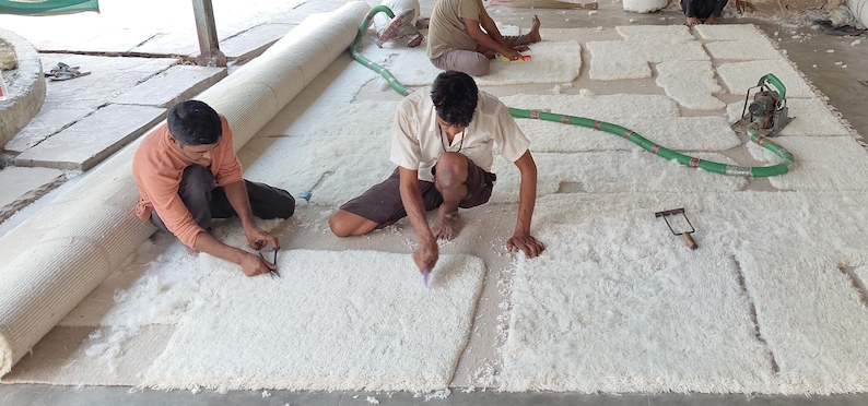 Puede incluir: Escena de taller donde se fabrica una alfombra grande de color blanquecino. Dos personas est&aacute;n arrodilladas, usando herramientas para trabajar en la superficie de la alfombra. Tambi&eacute;n se ve una alfombra enrollada y una manguera verde.