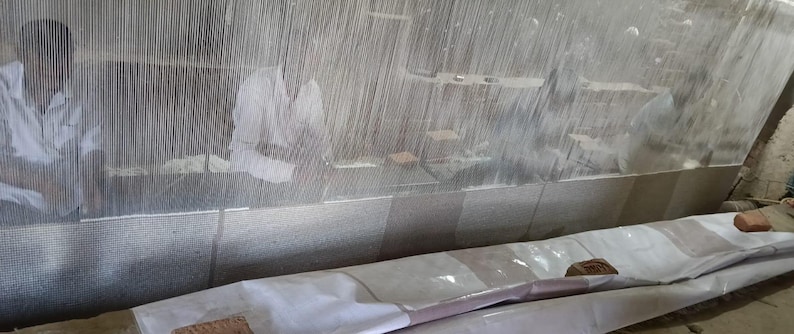Puede incluir: Taller textil con dos personas trabajando en un gran telar. El telar es de madera y metal, con hilos verticales estirados sobre el marco. Los trabajadores visten camisas blancas. El textil que se teje parece ser una alfombra.
