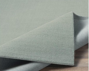 May include: A light blue rug with a textured surface, partially rolled up to reveal the underside. The rug's colour is a soft, muted blue-green, and the texture appears to be a looped pile. The rug is placed on a wooden floor.