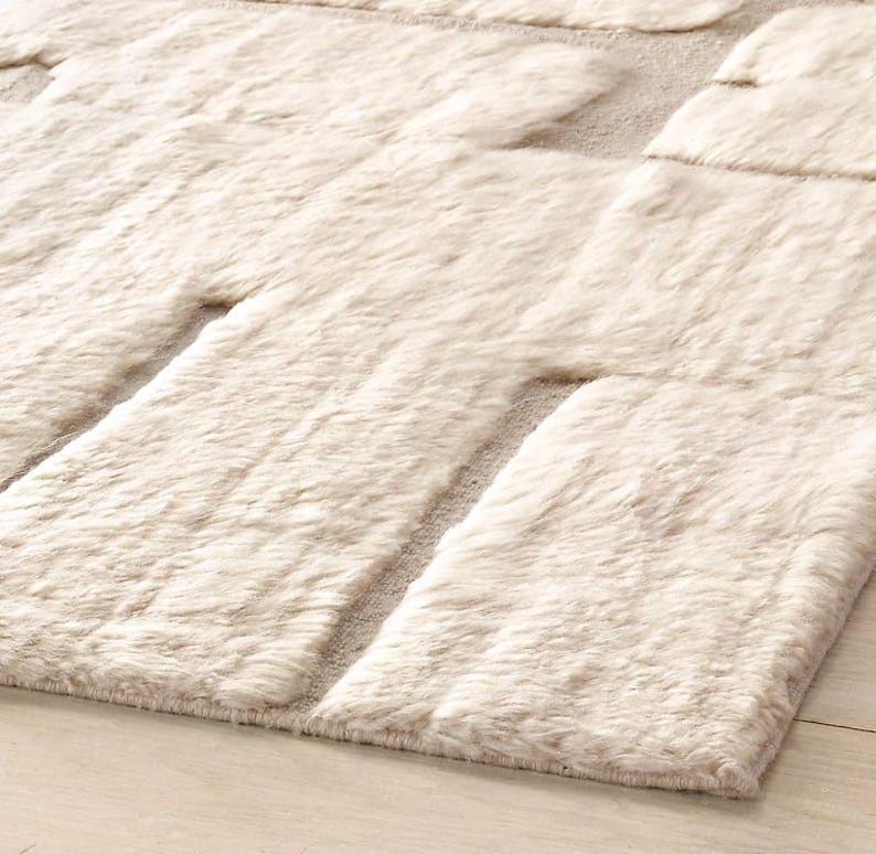 Puede incluir: Una alfombra rectangular de color crema con un patr&oacute;n geom&eacute;trico. La alfombra presenta secciones elevadas y texturizadas en un tono crema m&aacute;s claro, creando un aspecto afelpado y acogedor. La alfombra est&aacute; colocada sobre un suelo de madera clara.