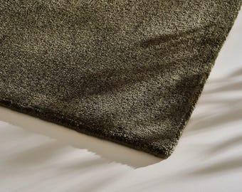 May include: Close-up of a section of a green rug. The rug has a dense, textured pile, and the color is a muted olive green. The corner of the rug is visible, showing the edge and the texture of the material. The rug is on a white surface.